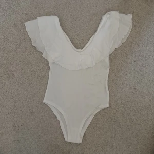 Vit offshoulder bodysuit med volang - Supersnygg vit bodysuit med bred volang runt axlarna och offshoulder-design. Perfekt för dig som vill ha en feminin och trendig look. Mjuk och stretchig känsla, passar till både jeans och kjol.