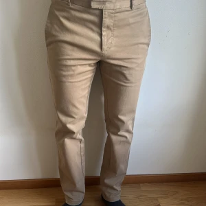Beige chinos från Tiger of Sweden - Snygga beige chinos från Tiger of Sweden i klassisk rak modell. Byxorna har bälteshällor, sidfickor och bakfickor med knapp. Tillverkade i bomull för en skön och stilren känsla. Perfekta för en clean och dressad look.