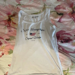 Vit linnetopp med New Hampshire-tryck - Vit linnetopp från Reaction med tryck 'New Hampshire Original Heritage U.S.A' och amerikansk flagga framtill. Klassisk ärmlös modell med rund halsringning, perfekt för en chill och avslappnad stil.