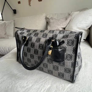 Loewe handväska med monogram - Säljer en snygg handväska från Loewe med klassiskt svart monogrammönster på grå/beige canvas. Väskan har svarta skinnhandtag, guldfärgade metalldetaljer och dragkedja. Perfekt storlek för att få plats med allt du behöver och riktigt stilren look.