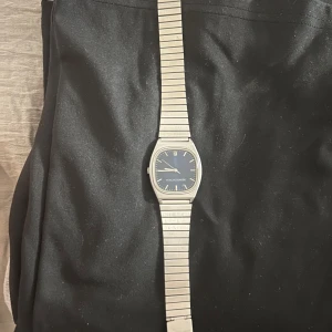 Silverfärgad klocka från Retroduction - Stilren klocka från Retroduction med silverfärgat armband i metall och mörkblå urtavla. Klockan har en rektangulär boett och tydliga indexmarkeringar. Perfekt accessoar för dig som gillar minimalistisk och klassisk design.