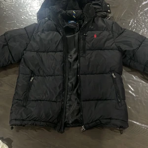 Svart pufferjacka från Polo Ralph Lauren - Säljer en svart pufferjacka från Polo Ralph Lauren med röd broderad logga på bröstet. Jackan har huva, flera dragkedjeförsedda fickor och quiltad design. Perfekt för kalla dagar och har en clean, sportig vibe.