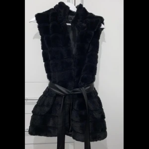 Svart pälsväst med bälte - Snygg svart pälsväst med markerad krage och ett brett, svart bälte i midjan. Västen har en fluffig och lyxig känsla med horisontella paneler och är perfekt för att lyfta vilken outfit som helst. Bältet är i skinnimitation och knyts i midjan för en cool siluett.