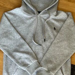 Grå hoodie från Polo Ralph Lauren - Snygg grå hoodie från Polo Ralph Lauren med klassisk huva och snörning. Tröjan har det ikoniska broderade logotypen på bröstet och är tillverkad i mjuk bomullsmix. Perfekt för en chill och stilren look.