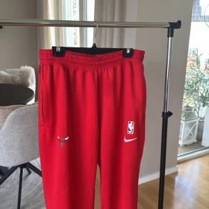 Röda Chicago Bulls mjukisbyxor Nike - Röda mjukisbyxor från Nike med Chicago Bulls-logga och NBA-märke. Byxorna har elastisk midja, dragkedja vid fickan och resår vid benslut. Perfekta för basketfans som vill ha en sportig och bekväm stil.