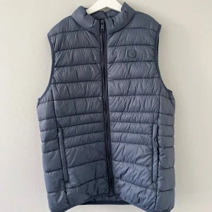 Grå / Blå - väst från Álvaro Moreno - Snygg grå dunväst från Alvaro Moreno med quiltad design och hög krage. Västen har dragkedja framtill, två fickor med dragkedja och en diskret logga på bröstet. Perfekt att slänga över en hoodie för extra värme. Västen är köpt i Spanien 🇪🇸- för ca 200/250 kr,      Inga defekter och använd endast fåtal gånger - hör av er vid frågor eller funderingar - Mvh Sigge🥂