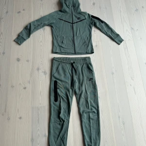 Nike grön träningsset med huva - Nike träningsset i dämpad grön färg med svart logga. Hoodien har dragkedja och huva framtill samt svarta detaljer på bröst och ärm. Byxorna har dragsko i midjan, svarta detaljer och dragkedjeficka på benet. Perfekt för dig som gillar sportig och avslappnad stil.