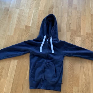Mörkblå hoodie Russedress XS - Mörkblå hoodie från Russedress i storlek XS. Tröjan har vit snörning i huvan, dragkedja framtill och två stora fickor. Mjuk insida och ribbade muddar vid ärmslut och nederkant. Perfekt för chill dagar eller när du vill vara extra bekväm.