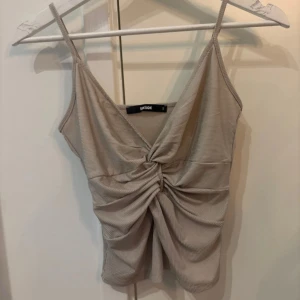 Beige ribbad linnetopp från Bik Bok - Supersnygg beige linnetopp från Bik Bok med smala axelband och twistad knut framtill. Toppen är ribbad och har en lätt glansig finish som ger en extra touch. Perfekt för dig som gillar stilrena och trendiga plagg med det lilla extra.