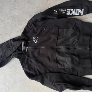 Svart Nike Air hoodie XS - Svart hoodie från Nike Air med dragkedja och huva. Stora fickor framtill, snörning i huvan och Nike Air-logga på bröstet samt ärmen. Skön och sportig stil, perfekt för chill eller träning.