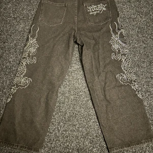 Ed Hardy jeans med drakmotiv - Snygga svarta jeans från Ed Hardy med feta vita drakmotiv längs båda benen och broderad logga på bakfickan. Raka ben och klassisk femficksmodell. Perfekta för dig som vill sticka ut med streetstyle och coola detaljer. Storlek 30/30
