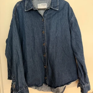 Oversized jeansskjorta från Selected Femme - Snygg oversized jeansskjorta från Selected Femme i klassisk blå denim. Skjortan har långa ärmar, knappar framtill och en avslappnad passform. Perfekt att bära öppen eller stängd för en cool och avslappnad look.