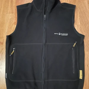 Svart Sail Racing fleeceväst - Svart fleeceväst från Sail Racing med Windstopper-teknologi. Västen har hel dragkedja framtill, två fickor med dragkedja och hög krage. Perfekt för lager-på-lager och outdoor-aktiviteter. Diskret logga på bröstet och nacken.