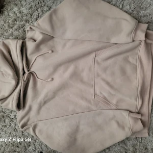 Beige hoodie med huva och ficka - Säljer en stilren beige hoodie med huva och dragsko. Tröjan har en stor magficka och ribbade muddar vid ärmslut och nederkant. Perfekt för en avslappnad look och enkel att matcha med det mesta.