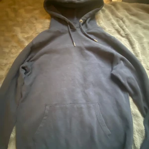 Mörkblå hoodie med huva och ficka - En stilren mörkblå hoodie med klassisk känguruficka framtill och justerbar snörning i huvan. Tröjan har långa ärmar och en relaxed passform, perfekt för chill dagar eller när du vill ha något bekvämt och enkelt.