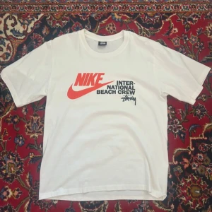 Vit Stüssy x Nike t-shirt L - Vit t-shirt från Stüssy i samarbete med Nike. Stor tryckt logga i rött och svart framtill med texten 'Nike International Beach Crew'. Klassisk passform och rund hals. Perfekt för dig som gillar streetwear och snygga prints.