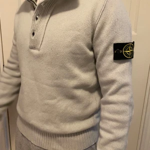 Ljusgrå stickad Stone Island tröja - Säljer en ljusgrå stickad tröja från Stone Island med klassisk patch på ärmen. Tröjan har hög krage med både dragkedja och knappar, samt ribbade muddar och nederkant. Perfekt för dig som gillar stilren och exklusiv streetwear. Vid snabb affär så kan eventuellt priset sänkas.