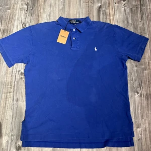 Blå pikétröja från Ralph Lauren - Klassisk blå pikétröja från Ralph Lauren med vit broderad logga på bröstet. Tröjan har korta ärmar, krage och två knappar framtill. Tillverkad i mjuk bomull som känns skön mot huden. Perfekt för en clean och stilren look.