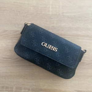 Svart axelväska från Guess - Snygg svart axelväska från Guess med guldigt logotyp på locket och diskret mönster över hela väskan. Väskan har en bred, justerbar axelrem och är tillverkad i syntetmaterial med struktur. Perfekt storlek för det viktigaste och stilren design. Aldrig använd (pris kan diskuteras )💕