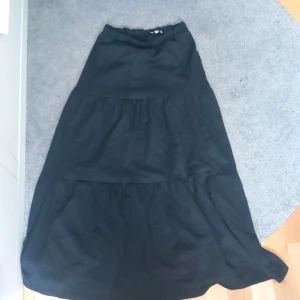 Svart långkjol från Samsa Young - Svart långkjol från Samsa Young med volangdetaljer och resår i midjan. Kjolen har en luftig och flowy passform som gör den superbekväm och lätt att styla. Perfekt för dig som gillar en enkel men snygg look.