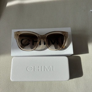 Beige solglasögon från CHIMI - Snygga solglasögon från CHIMI i transparent beige med bruna tonade glas. Modellen har en trendig, något cat eye-inspirerad form och tjockare bågar i plast. Perfekta för att lyfta din stil under soliga dagar. Levereras med originaletui. Solglasögonen är oanvända. Köparen står för frakt.