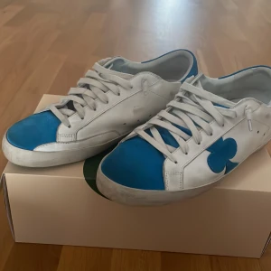 Vita och blå sneakers från The Gallerist - Snygga vita sneakers med blå detaljer och klubb-symbol på sidan från The Gallerist. Skorna har snörning, platt sula och är tillverkade i skinn. Perfekta för dig som gillar unik design och vill sticka ut med coola detaljer.