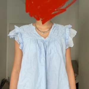 Ljusblå blus med volangärmar - Supersöt ljusblå blus med v-ringning och volangdetaljer på ärmarna. Blusen har broderade partier upptill och är tillverkad i ett luftigt material, perfekt för varma dagar. Den är ärmlös och har en lös passform som ger en chill vibe.