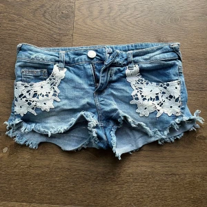 Jeansshorts med spets från Mossimo - Blå jeansshorts från Mossimo med fransig kant och vita blommiga spetsdetaljer framtill. Klassisk femficksmodell, lågmidjade och i mjuk bomull med lite stretch. Perfekta för sommaren och ger en chill vibe.