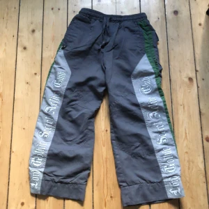 Grå baggy byxor med tryck - Baggy grå byxor med bred passform och gröna detaljer längs sidorna. Stora silvriga paneler med texten 'Loud Gothic Street Active Club' på båda benen. Resår och snörning i midjan samt fickor bak med lock. Perfekta för en streetwear-look.