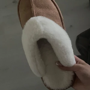 Bruna tofflor med vit fuskpäls - Mysiga bruna tofflor med fluffig vit fuskpäls runt öppningen och insidan. Utsidan är i mockaliknande material och sulan är vit och räfflad för extra grepp. Storlek 36/37. 