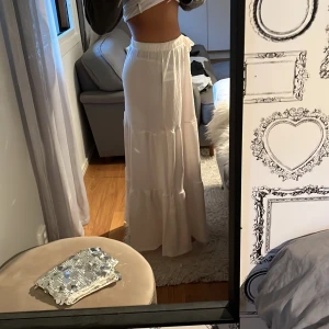 Vit långkjol med volanger från 157 - Superfin vit långkjol från 157 med volanger och resår i midjan. Kjolen har en snygg knytning framtill och är gjord i ett lätt, flowy tyg som känns perfekt för varmare dagar. Passar dig som gillar en chill och bohemisk stil.