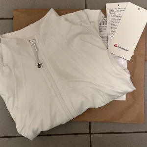 Vit crop jacka från Lululemon - Snygg vit crop jacka från Lululemon med figurnära passform och dragkedja framtill. Jackan har långa ärmar, hög krage och två fickor med dragkedja. Perfekt för träning eller chill dagar. Materialet är mjukt och stretchigt för maximal komfort. Har använt en gång. Ord: 1280 och säljs nu för 800-850kr. Skriv för att diskutera pris😊
