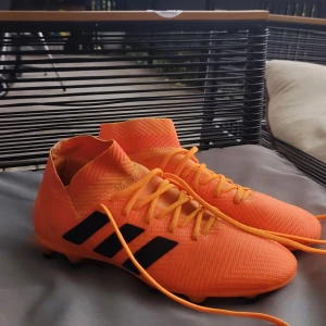 Adidas Nemesis orange fotbollsskor - Säljer ett par Adidas Nemesis fotbollsskor i en riktigt snygg och iögonfallande orange färg med svarta detaljer. Skorna har snörning, högre skaft och är tillverkade i syntetmaterial med mönstrad yta för extra grepp på planen.