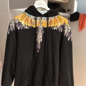 Marcelo Burlon hoodie med fjädermönster - Svart hoodie från Marcelo Burlon County of Milan med ikoniskt fjädermönster i gult, orange, blått och vitt över axlar och bröst. Tröjan har huva med snörning och stor känguruficka framtill. Tillverkad i 100% bomull för en skön och avslappnad vibe.