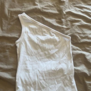 Vit oneshoulder topp från Stories XS - Säljer en vit ribbad oneshoulder topp från Stories i storlek XS. Toppen är ärmlös med asymmetrisk axel och har en tight passform. Perfekt för dig som gillar minimalistisk och stilren look.aldrig använd!!