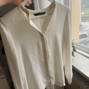 Vit blus med knappar från Zara Basic - Säljer en stilren vit blus från Zara Basic i storlek S. Blusen har v-ringning, knappar framtill och långa ärmar. Baksidan har snygga plisserade detaljer som ger en extra touch. Materialet känns lätt och flowy, passar perfekt under en cardigan.