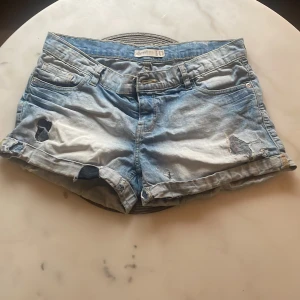 Ljusblå slitna lågmidjade jeansshorts Denim  - Ljusblå lågmidjade jeansshorts från Denim  med slitna detaljer och hål för en cool, avslappnad look. Klassisk femficksmodell, uppvikta benslut och knappgylf. Perfekta för varma dagar när du vill ha en chill vibe. midjemått 41 cm