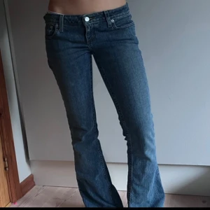 Blå bootcut lågmidje jeans med fickdetalj - Snygga blå bootcut jeans med låg midja och coola fickdetaljer bak, inklusive knapp med graverad logga. Jeansen har klassisk femficksdesign och är gjorda i ett mjukt denimtyg som sitter skönt. Perfekta för dig som gillar en avslappnad men trendig look.