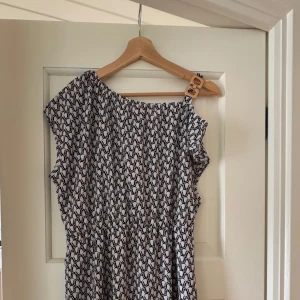 Svartvit mönstrad klänning - Snygg svartvit klänning med grafiskt mönster och en cool detalj med spänne på ena axeln. Klänningen har en avslappnad passform och är ärmlös, perfekt för varma dagar. Materialet känns lätt och luftigt.