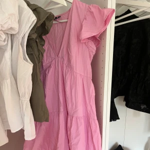 Rosa volangklänning med v-ringning - Supersöt rosa klänning med v-ringning och volangärmar. Den har en luftig passform och är sydd i flera lager för extra volym. Perfekt för dig som gillar en feminin och trendig stil.