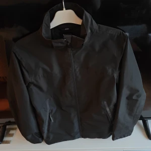 Svart vindjacka från Everest 158/164 - Svart vindjacka från Everest med hög krage och dragkedja framtill. Jackan har två fickor med dragkedja och en diskret reflexdetalj på ryggen. Perfekt för blåsiga dagar och enkel att matcha med allt. Snygg och funktionell för dig som gillar stilren look.