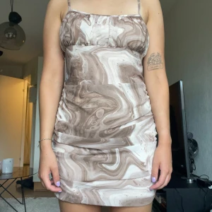 Brun mönstrad kortklänning med smala band - Snygg kortklänning med smala axelband (som man knyter själv) och marmorerat mönster i olika nyanser av brunt och beige. Klänningen har en figurnära passform och draperad effekt nertill. Aldrig använd, prislapp kvar 