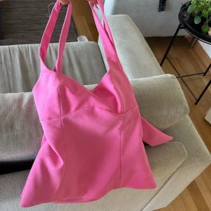 Topp  med rosett från Zara - Supersöt axelväska från Zara i starkt rosa färg. Väskan har en unik form med breda axelband och en stor rosett på sidan som ger den en extra touch. Tillverkad i ett slätt syntetmaterial som känns mjukt och följsamt. Perfekt för dig som vill sticka ut med en färgglad accessoar. Storlek Xs.  Pris kan diskuteras