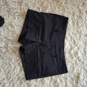 Svarta klassiska shorts med fickor - Snygga svarta shorts med klassisk look och raka ben. De har två fickor fram och två bak, samt hällor för bälte. Perfekta för sommaren och passar till allt. Materialet känns som bomull eller polyester och de har en stilren, enkel design.