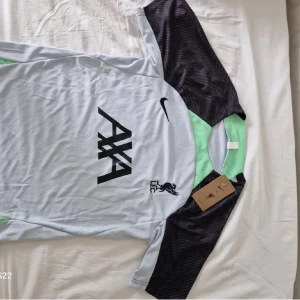 Liverpool träningsset Nike S - Liverpool FC träningsset från Nike i storlek S. Tröjan är ljusgrå med svarta ärmar och mintgröna detaljer, samt stor AXA-logga och LFC-märke på bröstet. Matchande svarta shorts med mintgröna inslag och klubbmärke. Tillverkad i lätt och ventilerande polyester.
