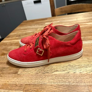 Novita Amira sneakers  - Snygga röda sneakers från Novita i mjuk mocka med vita sulor. Skorna har röda snören, guldfärgade detaljer och spänne samt en metallbricka med Novita-logga. Perfekt för dig som vill sticka ut med färg och stil. Nypris 1400kr. 