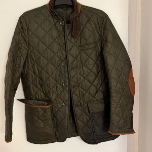 Quiltad jacka från Oscar Jacobson - Mörkgrön quiltad jacka från Oscar Jacobson med bruna detaljer på armbågar, krage och fickor. Klassisk design med knappar framtill, flera fickor och snyggt rutmönster. Perfekt för dig som gillar stilren och tidlös look.