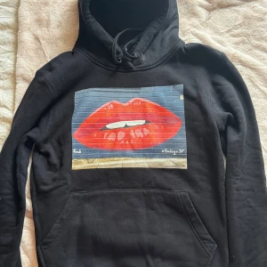 Svart hoodie The Cool Elephant Lips - Svart hoodie från The Cool Elephant med stor grafisk print av röda läppar på bröstet. Klassisk huva med snörning och känguruficka framtill. Mjuk och skön bomullsblandning, perfekt för en chill och trendig look. I storlek S och superfint skick! Pris kan diskuteras!