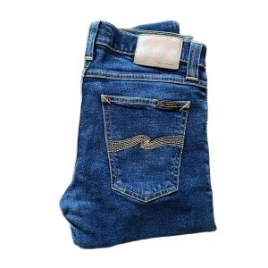 Nudie Jeans Tight Long John blå jeans - Snygga blå jeans från Nudie Jeans, modell Tight Long John. De har klassisk femficksdesign, kontrastsömmar och smal passform. Tillverkade i ekologisk bomull med lite stretch för extra komfort. Perfekta för dig som gillar en tight och stilren look. Midja ca 36cm se liten defekt på högerknäet