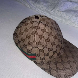 Gucci keps med GG-mönster och läderdetalj - Snygg keps från Gucci i beige och brunt GG-monogram med klassiska gröna och röda ränder på sidan. Justerbar rem i brunt läder baktill och insida i mjukt brunt tyg. Perfekt för dig som vill sticka ut med en lyxig accessoar.